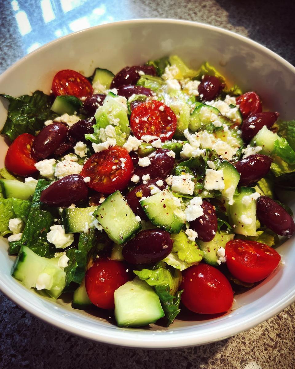 Ensalada fresca con tomate cherry, pepino, aceitunas negras, lechuga y queso feta en un bowl blanco.