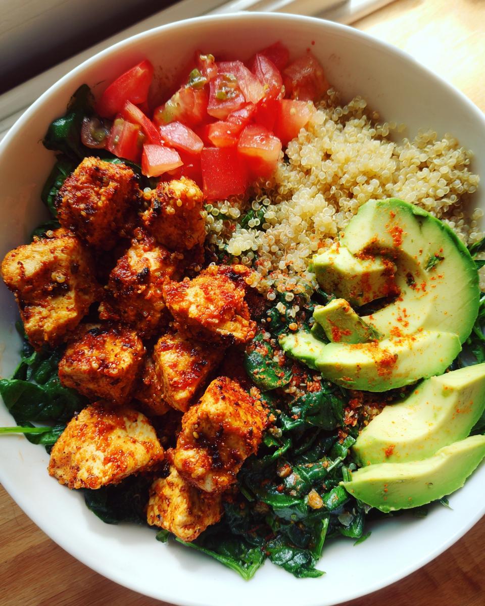 Bowl saludable de pollo y quinoa con espinacas, aguacate y tomate fresco en un plato blanco.