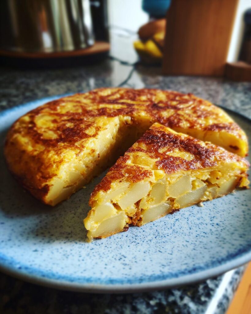 Tortilla de patatas tradicional con una porción cortada en un plato azul, receta de tapas españolas.