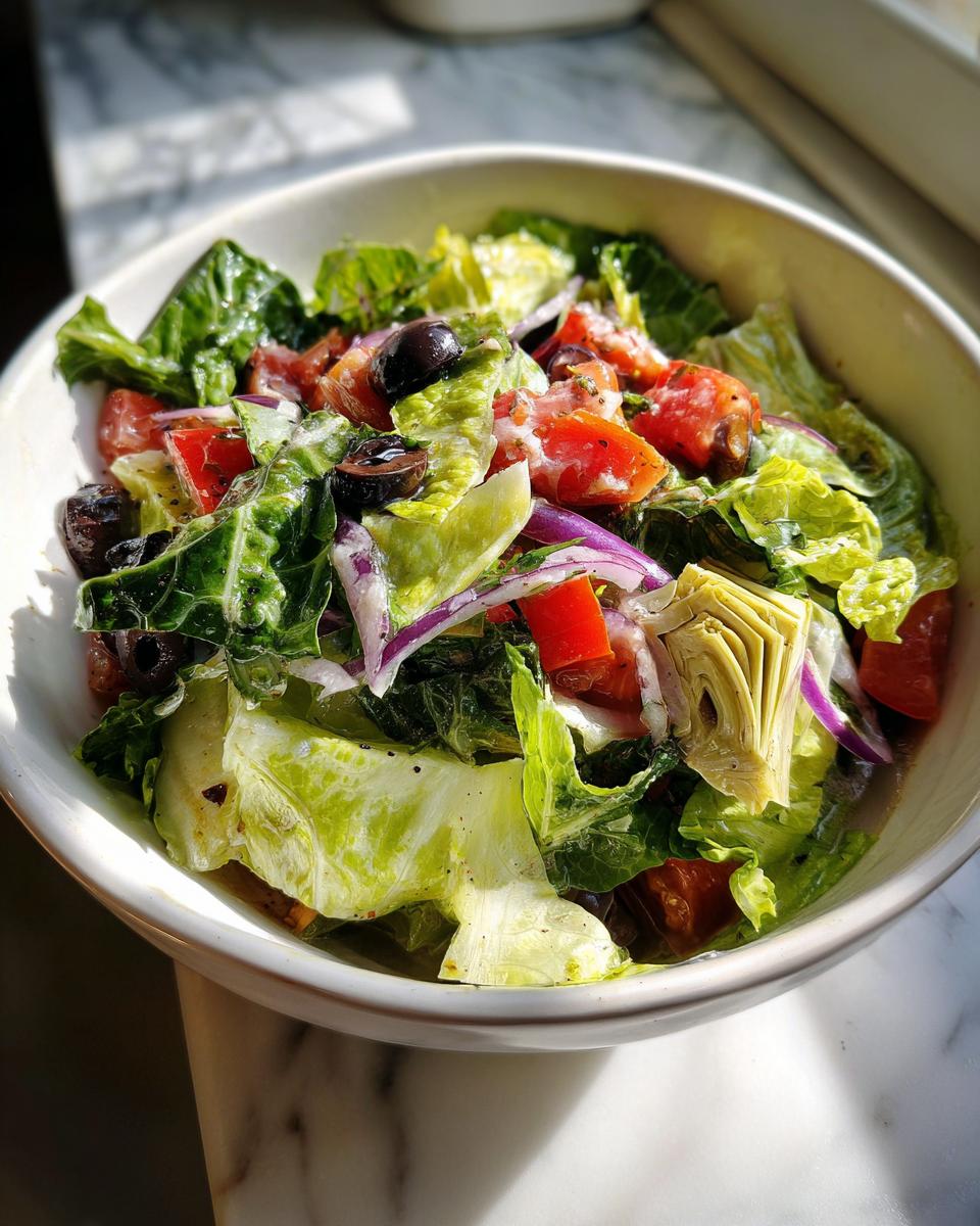 Ensalada con alcachofas, lechuga, tomate, aceitunas negras y cebolla morada en un bol blanco.